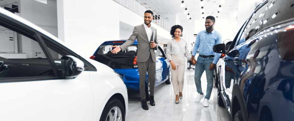Car Selling Business. Manager Showing Luxury Automobile To Afro Spouses In Automobile Dealership Center. Panorama