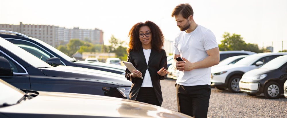 Car saleswoman dressed smartly holding a tablet, showing a man in a white top and black trousers a car within the dealership.